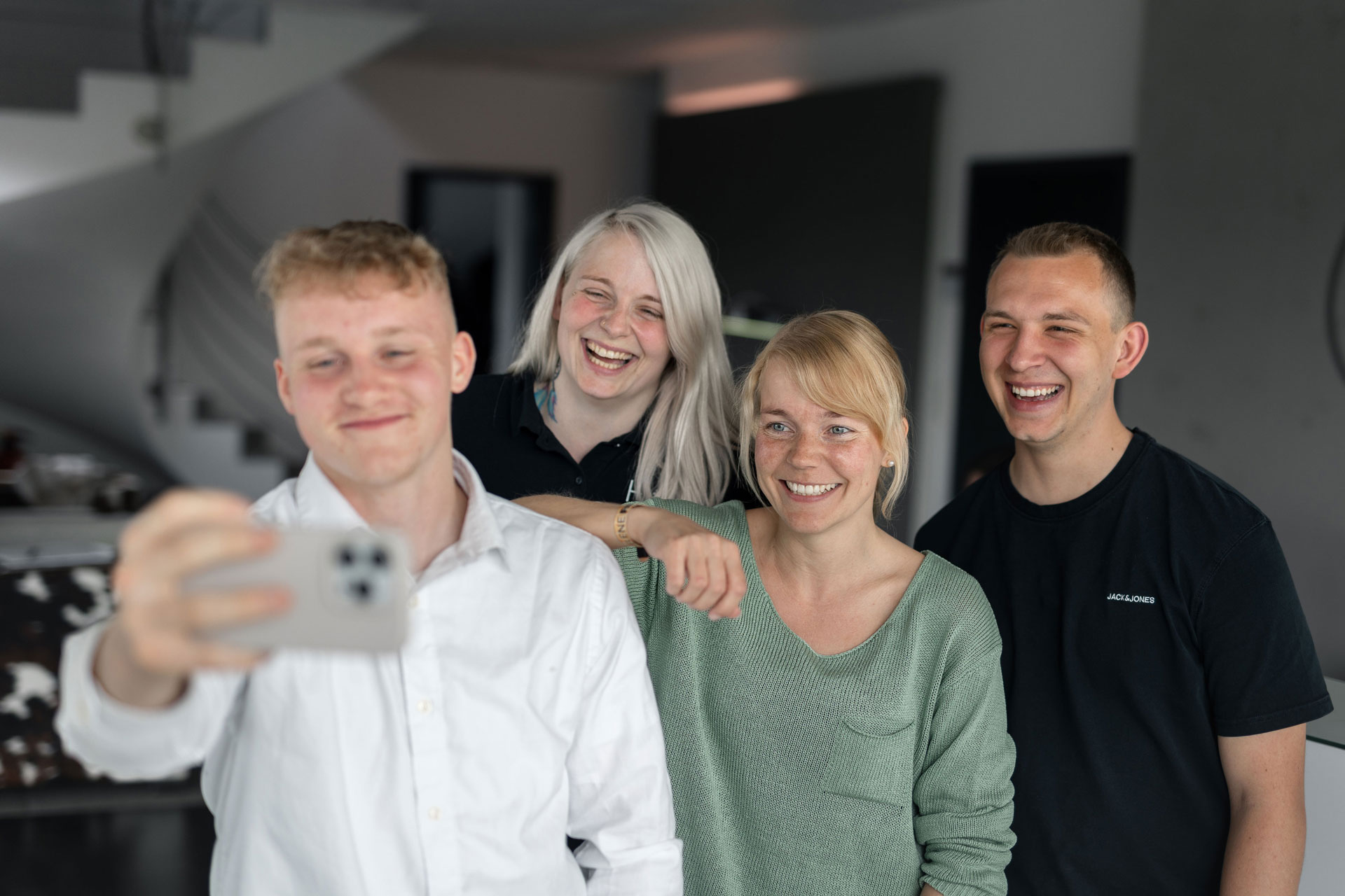 Vier junge Menschen (2 Männer und 2 Frauen) um die 20 Jahre machen ein Selfie. Der vordere Mitarbeiter hält dabei das Smartphone. Eine der jungen Frauen lehnt lässig den Arm auf seiner Schulter ab. Alle lächeln fröhlich in die Kamera. Sie sind in den Farben schwarz, weiß und mintgrün gekleidet und geben somit ein stimmiges Bild ab.