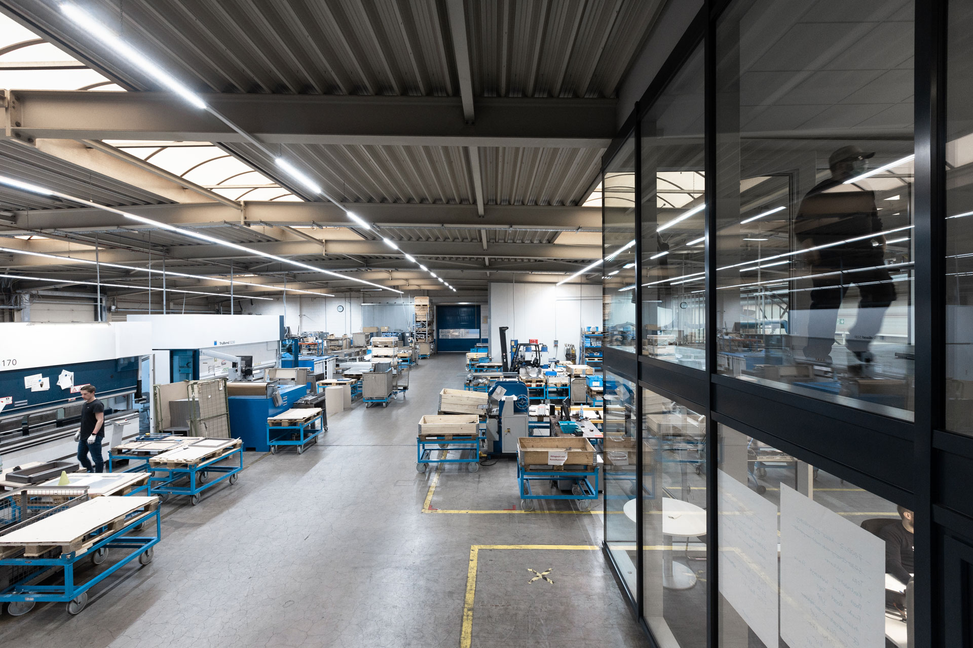 Drohnenaufnahme von einem Teil einer Fertigungshalle. Maschinen mit davorstehenden Paletten und Mitarbeitern. Im rechten Bildrand wird ein Konstruktionsbüro aus Glas angeschnitten.